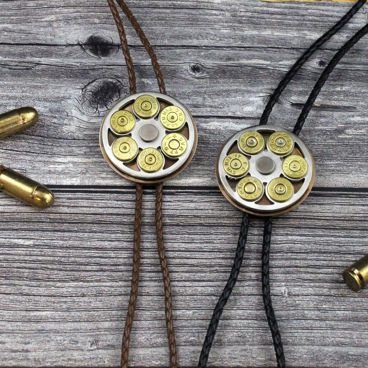 Two shotgun shell bolo ties displayed on a weathered gray wood background. One tie is brown with a silver-toned metal disc surrounded by brass-colored bullet casings, and the other is black with a matching silver-toned metal disc.