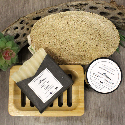 A styled product arrangement on a gray wood surface featuring a bar of whipped tallow soap in a dark gray wrapper, a round natural loofah bath sponge, and a small jar labeled whipped tallow, with a bamboo soap dish and decorative props around them.