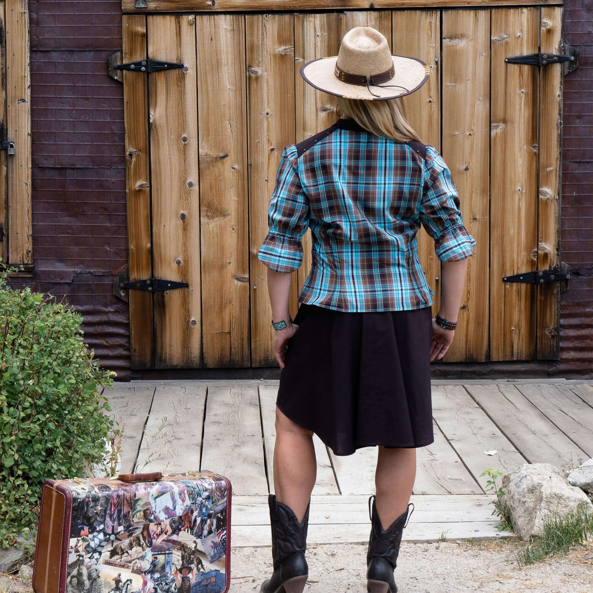 Woman wearing a Two-Step Women's Western Shirt with plaid pattern, cowboy hat, and boots, standing in front of a wooden barn.