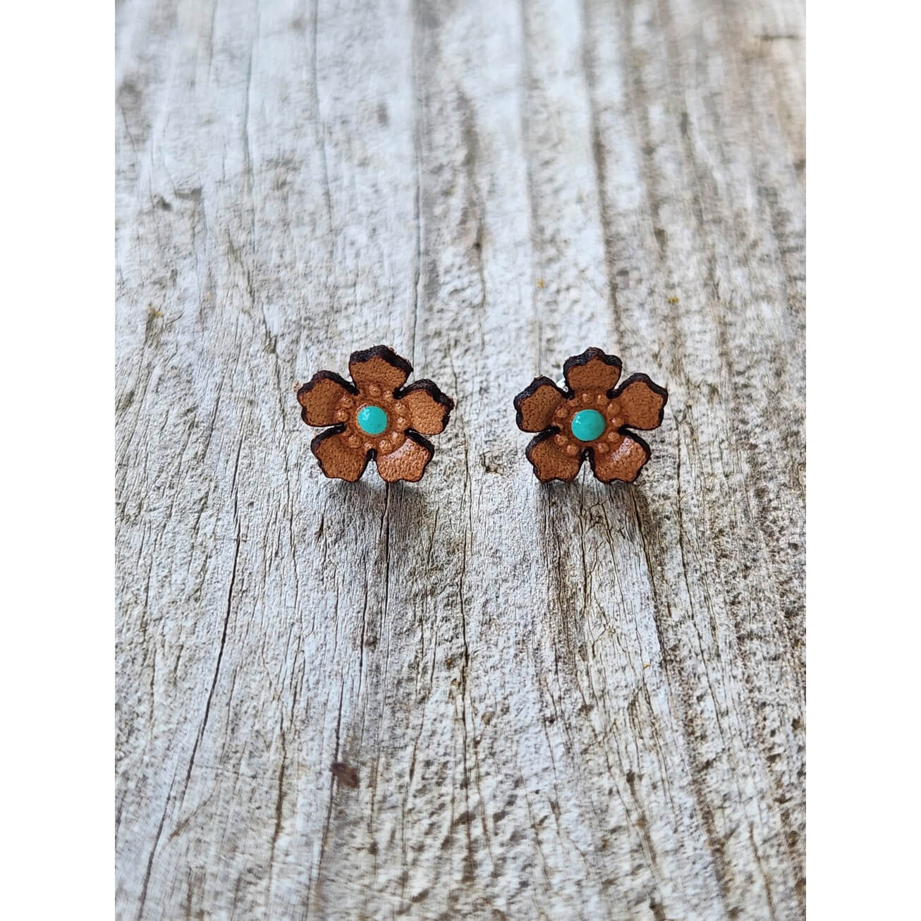 A pair of small flower-shaped stud earrings with brown petals and turquoise centers, displayed on a weathered light wood surface.
