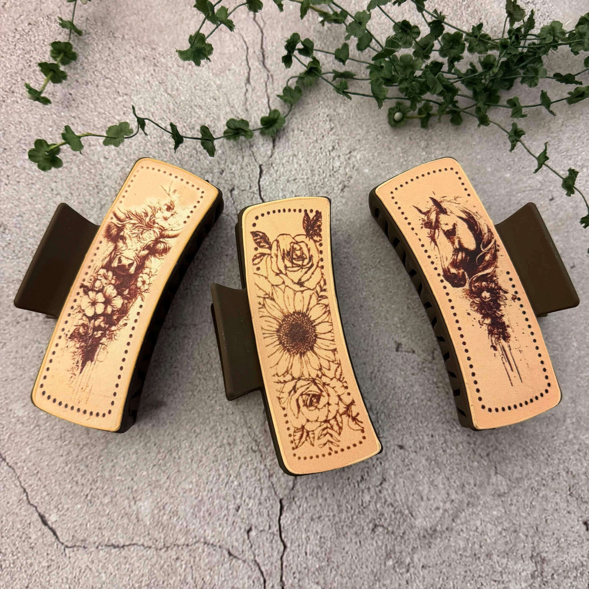 Three hair clips with designs on a textured surface with greenery.
