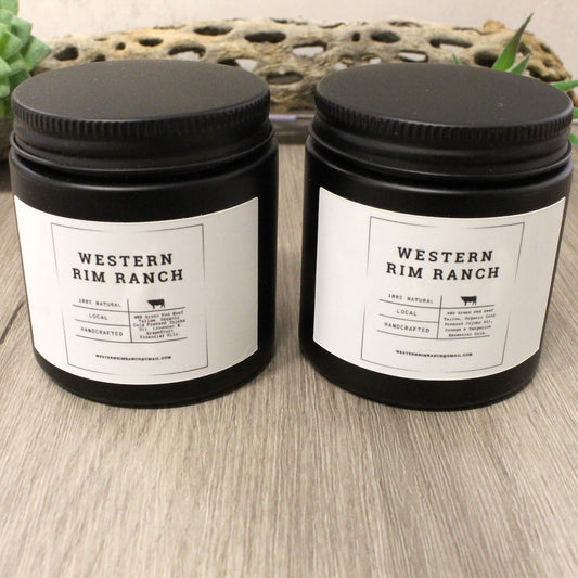 Two black plastic jar candles with white labels reading 'WESTERN RIM RANCH' sit on a light wood surface in front of a woven decorative mat.