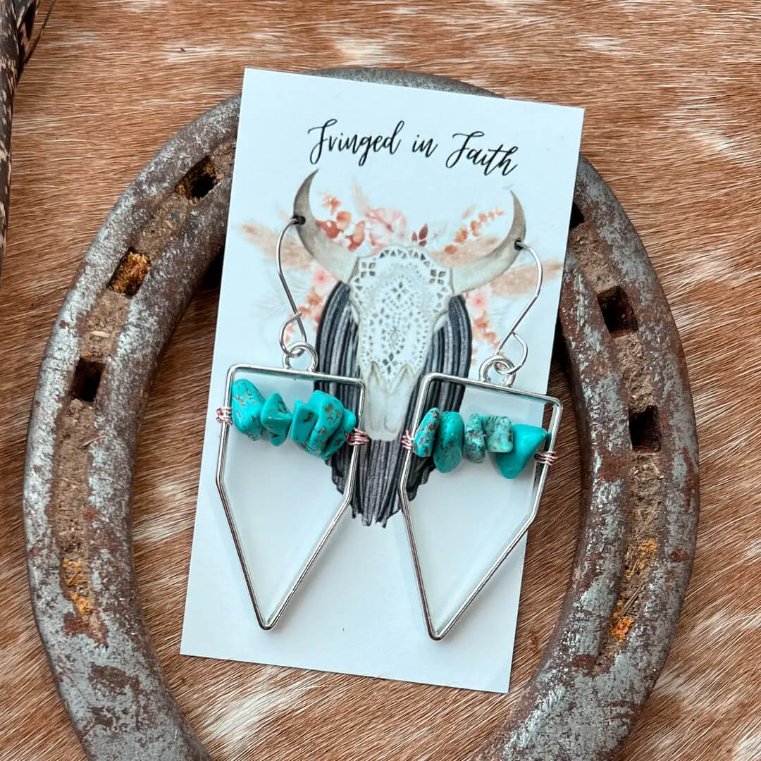 A pair of bohemian-style dangle earrings displayed on a white card labeled 'Fringed in Faith.' The earrings feature silver-toned metal frames, small black-and-white striped beads, and bright turquoise-colored stones, resting on a rustic metal horseshoe over a brown fur surface.
