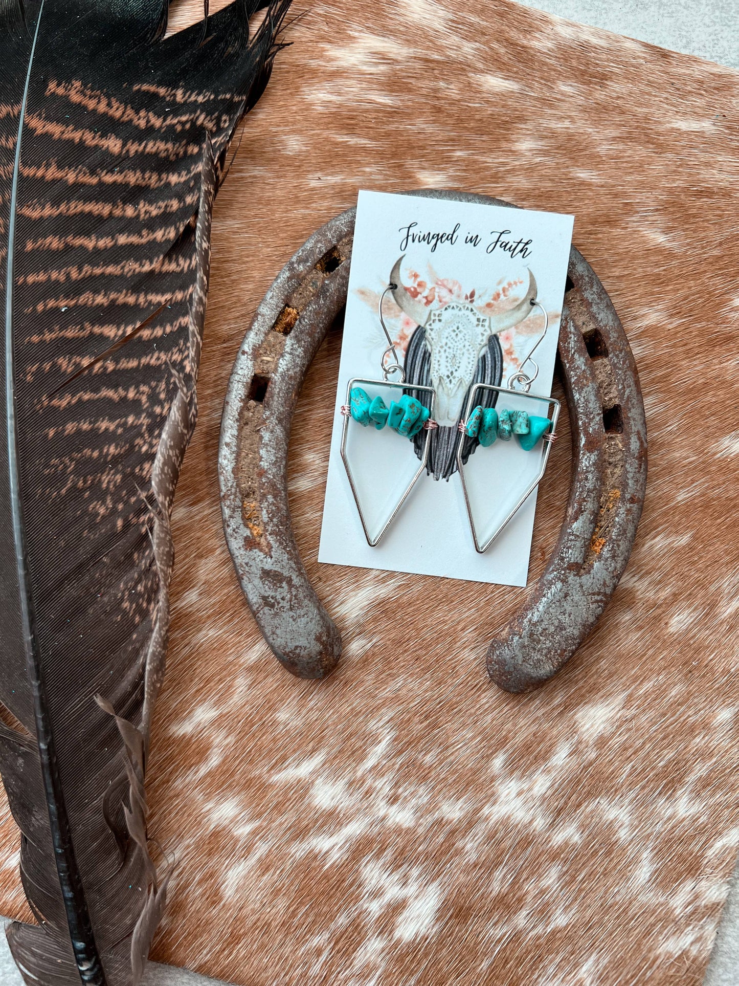 A pair of silver-toned dangle earrings with turquoise beads. The card is placed on a rustic brown and white hide surface next to a large feather and a weathered horseshoe.