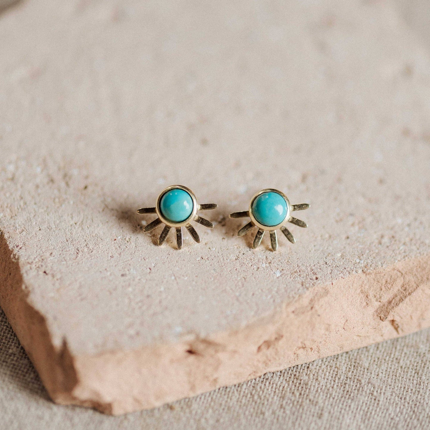 A pair of gold-toned sunburst stud earrings with round turquoise blue centers, displayed on a textured beige stone slab.