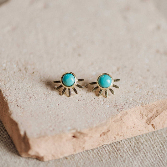 A pair of gold-toned sunburst stud earrings with round turquoise blue centers, displayed on a textured beige stone slab.