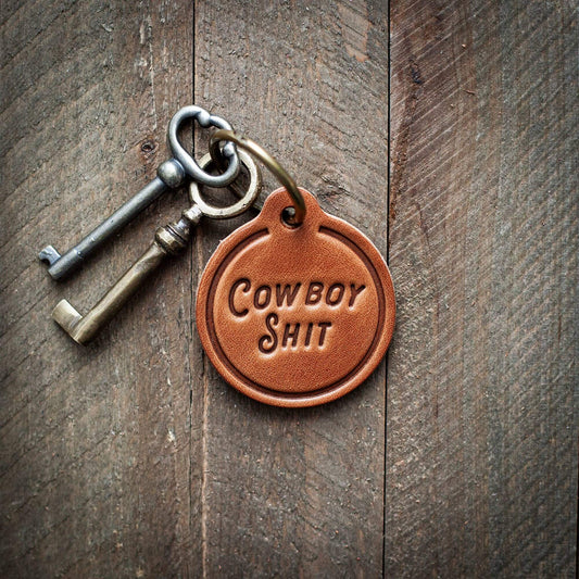 Cowboy Shit Leather Keychain featuring a classic design on a wooden surface.