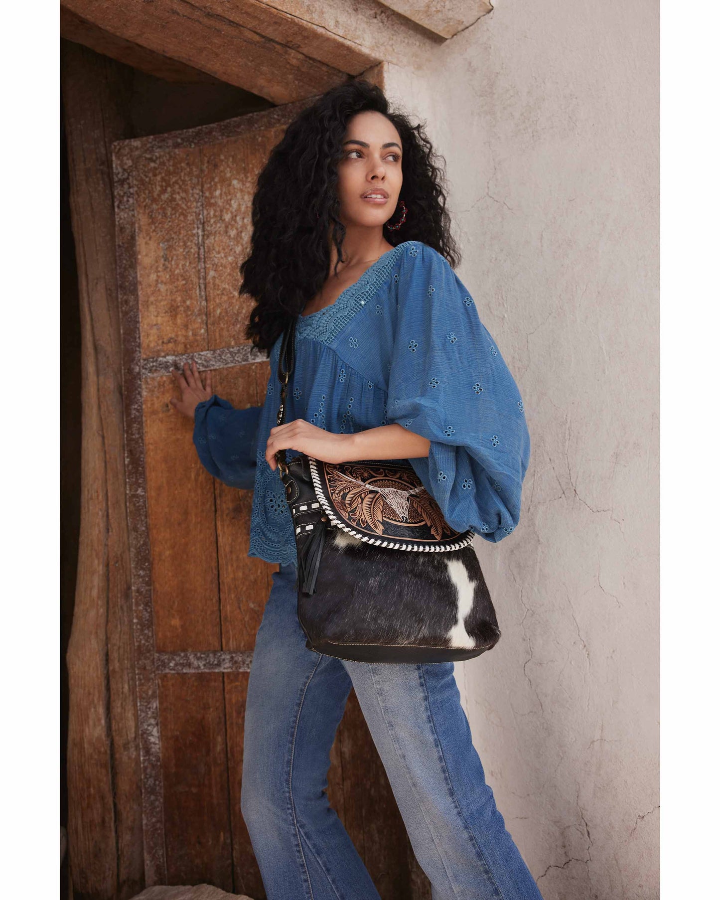 Woman holding a black handbag with intricate designs, wearing a blue top and jeans, against a rustic wooden door.