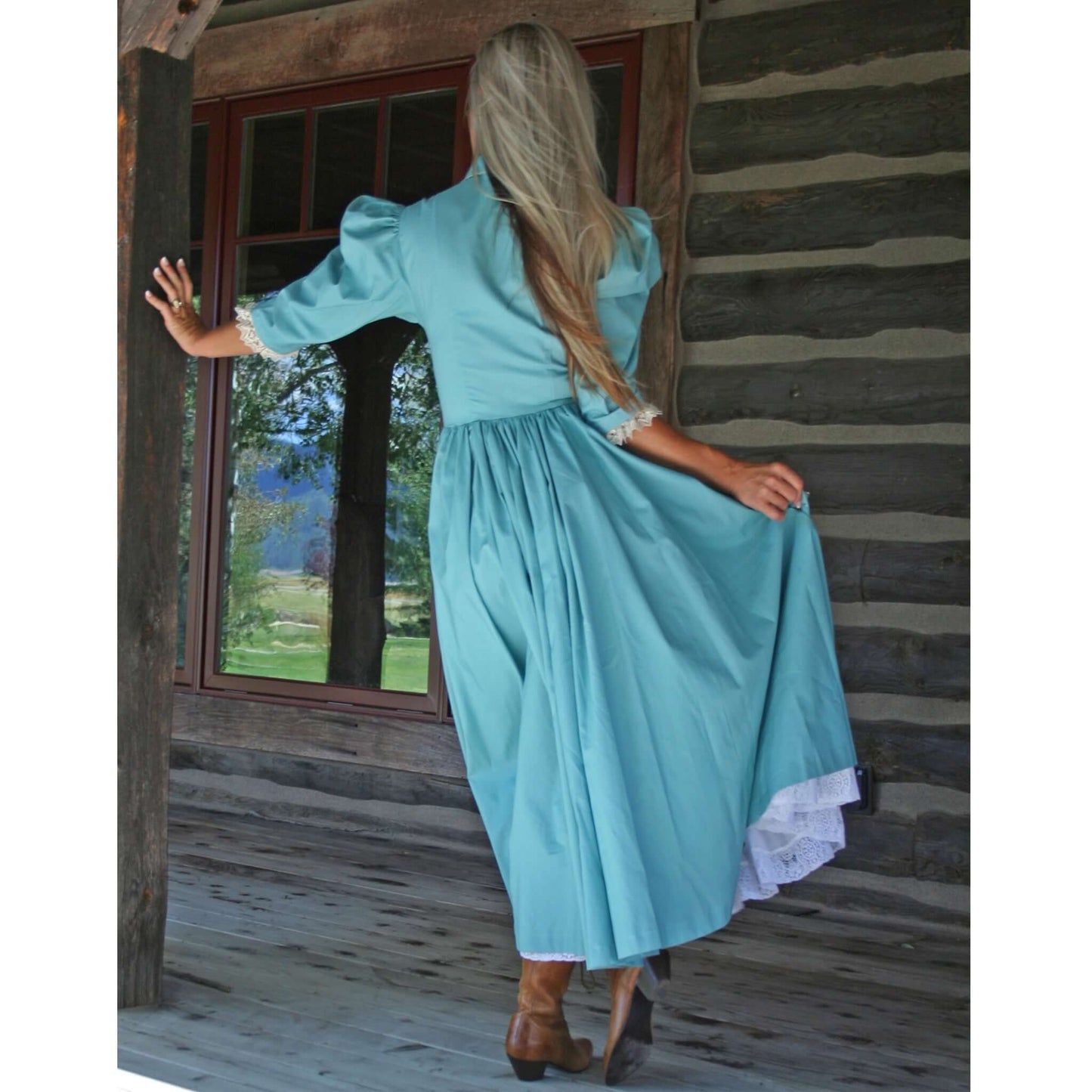 Woman in a frontier homestead western dress, showcasing ¾ sleeves and full skirt, standing on porch with scenic background.