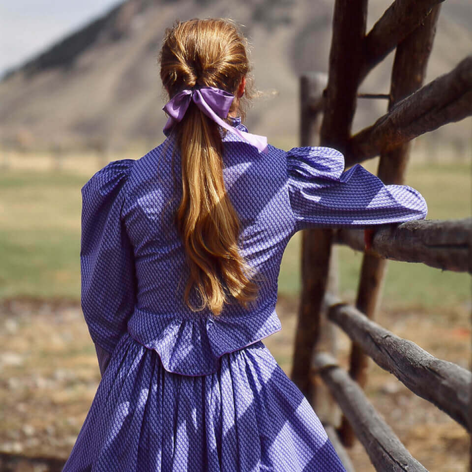 Back view of a woman in a vintage purple Old West outfit, showcasing puffed sleeves and a flowing skirt with a rustic backdrop.